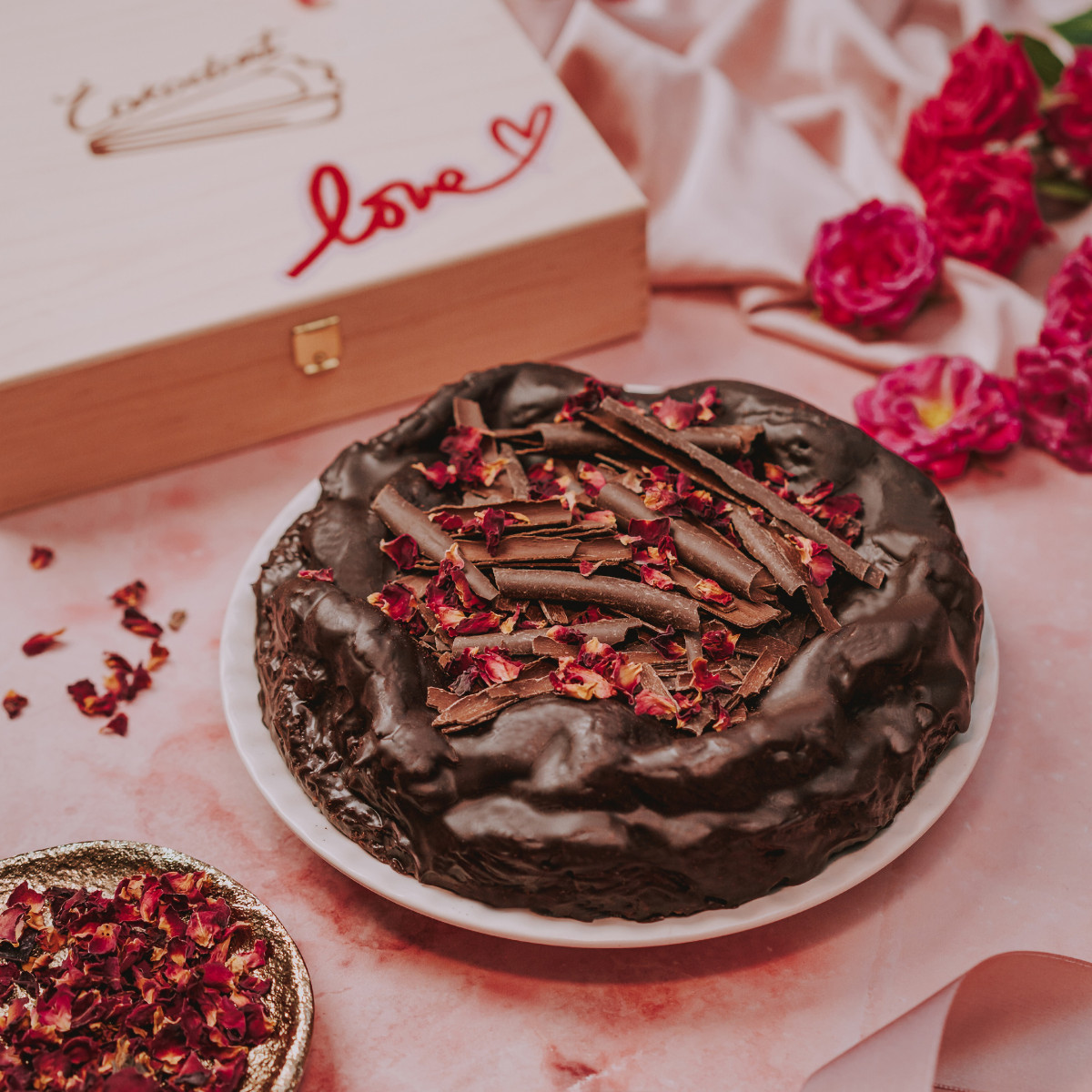 Valentýnský čokoládový dort s lyofilizovanými růžemi vedle dřevěné dárkové krabice s nápisem Love a rozptýlenými růžovými poupěty na růžovém podkladu