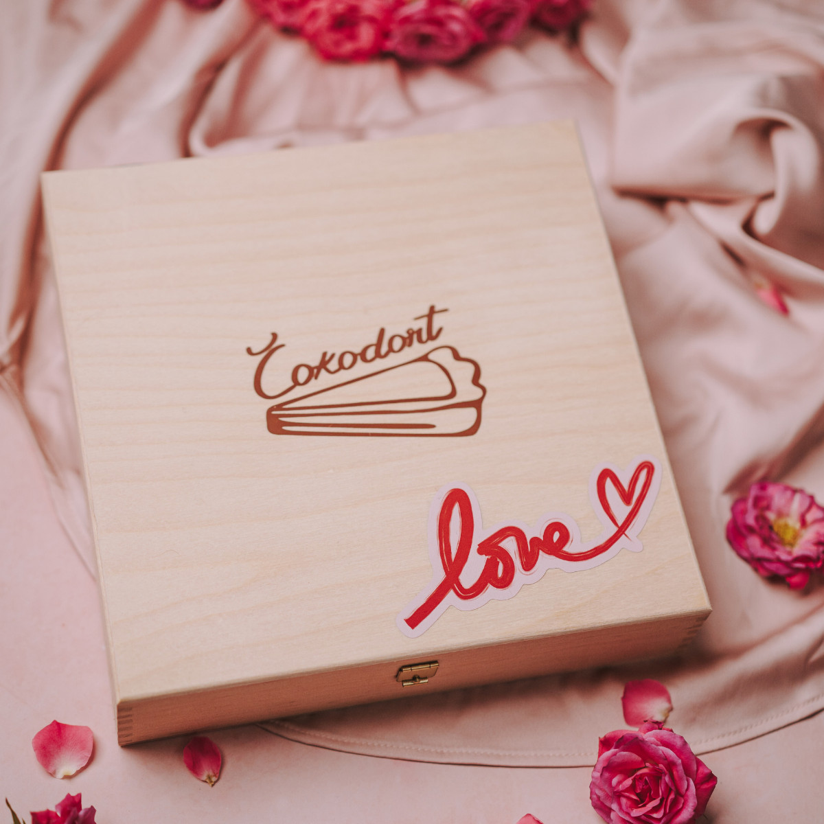 Dřevěná dárková krabice Čokodort s elegantním logem a nápisem "Love" v srdcovém designu, obklopená romantickými růžovými poupěty na saténové růžové látce